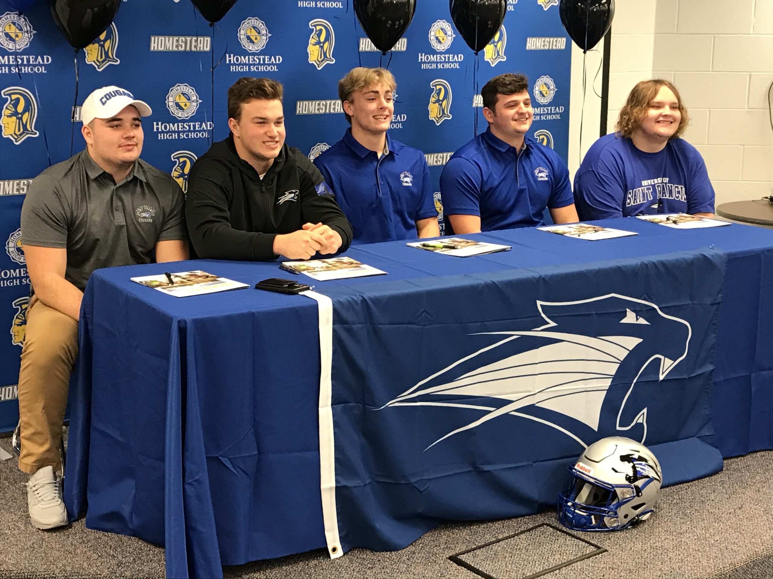 Five young men seated at a table with a blue tablecloth featuring a sports team logo.