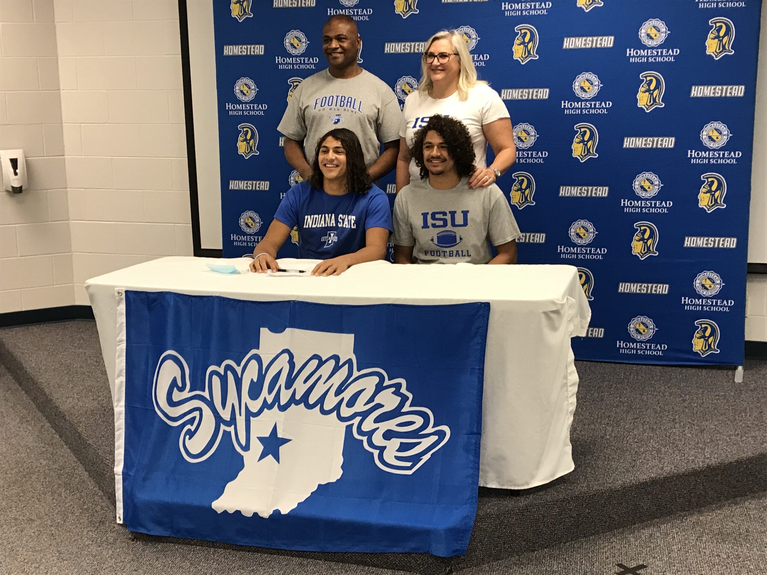 Two students signing documents with supportive people behind them.