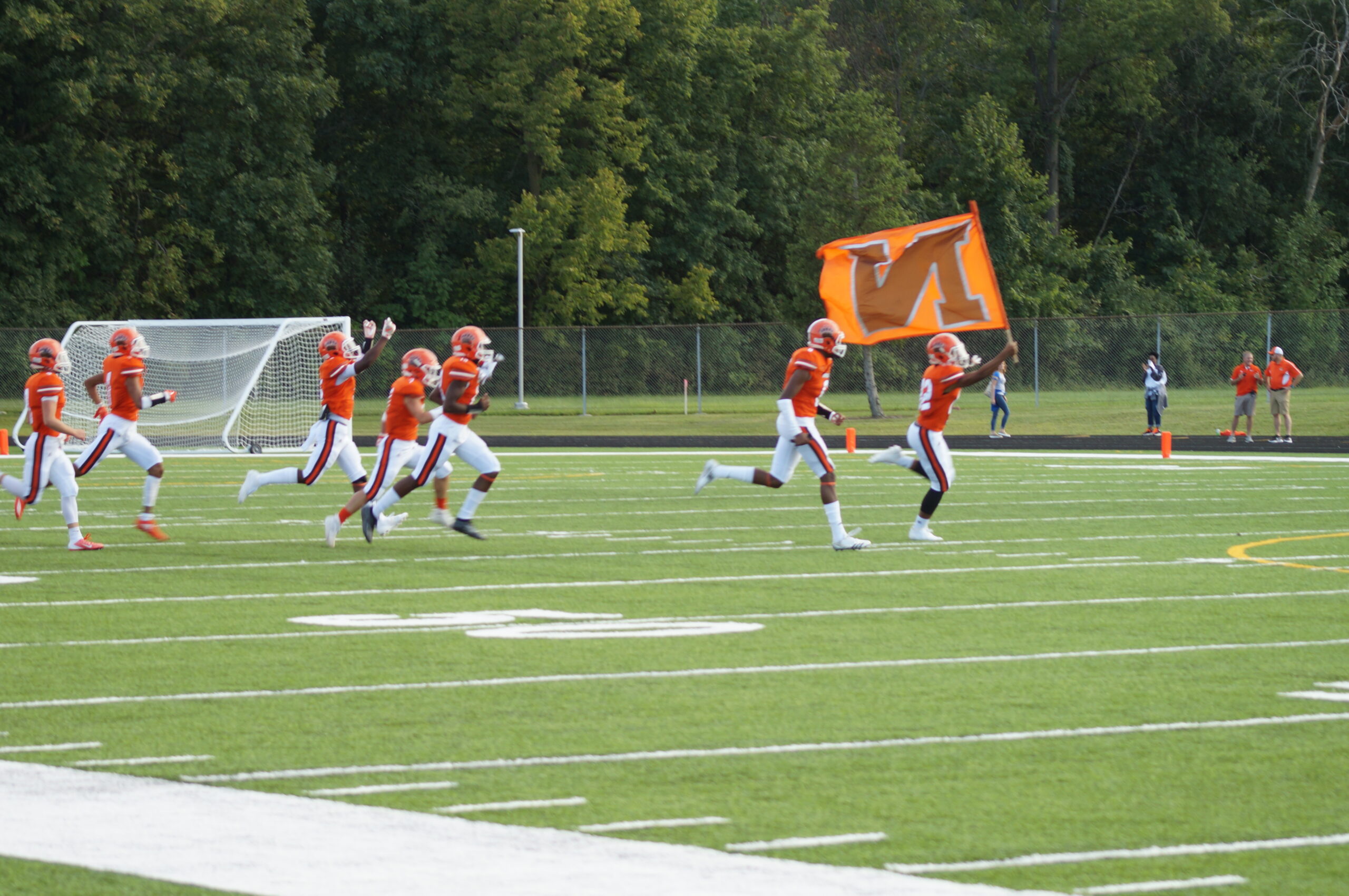 Young athletes running on a football field, one holding an orange flag.