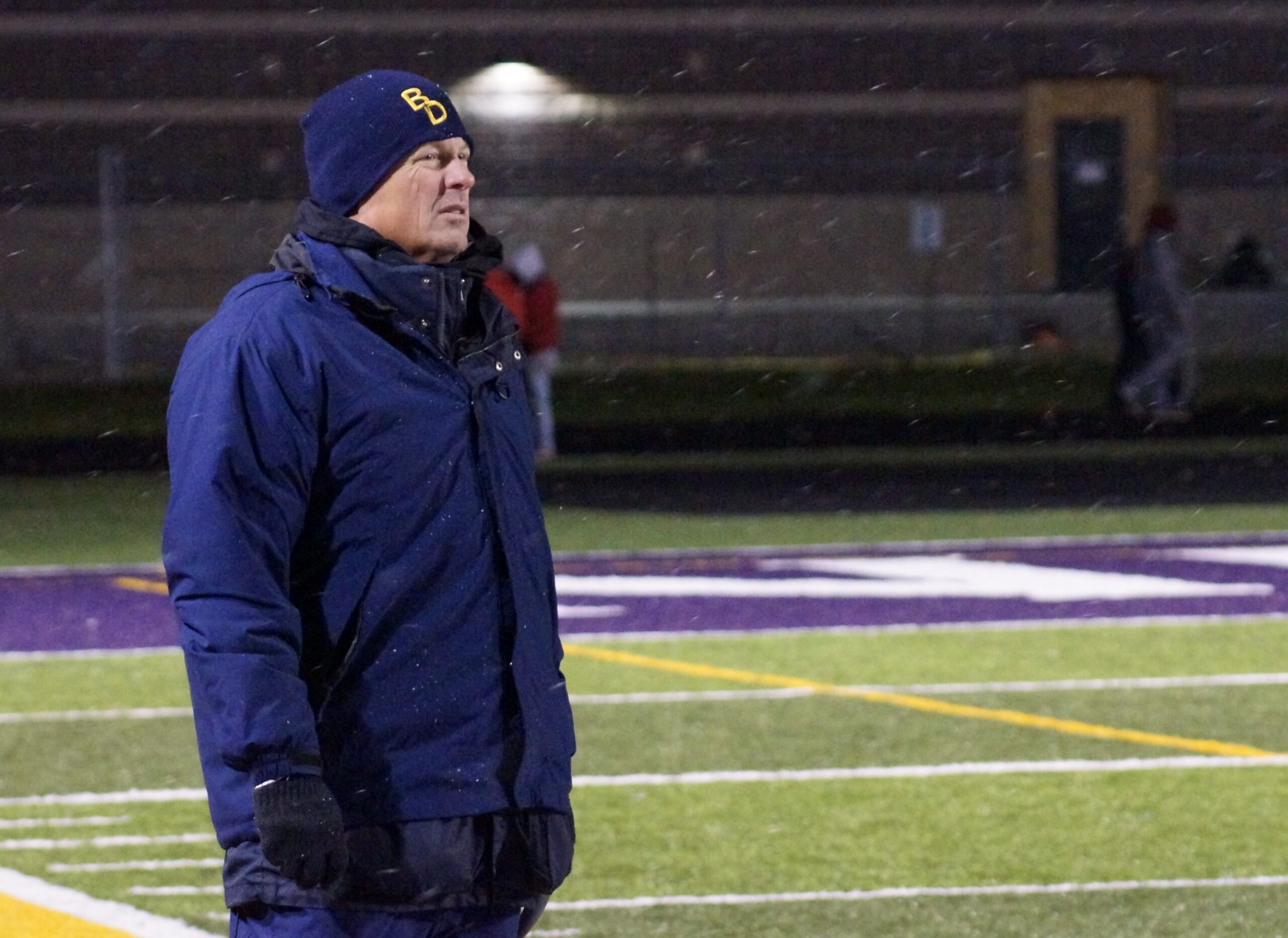Person in blue winter coat and hat standing on a sports field at night.